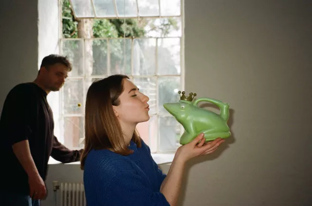 junge frau hält eine grüne figur in der hand, die ausschaut wie ein frosch und tut so als würde sie die figur küssen.