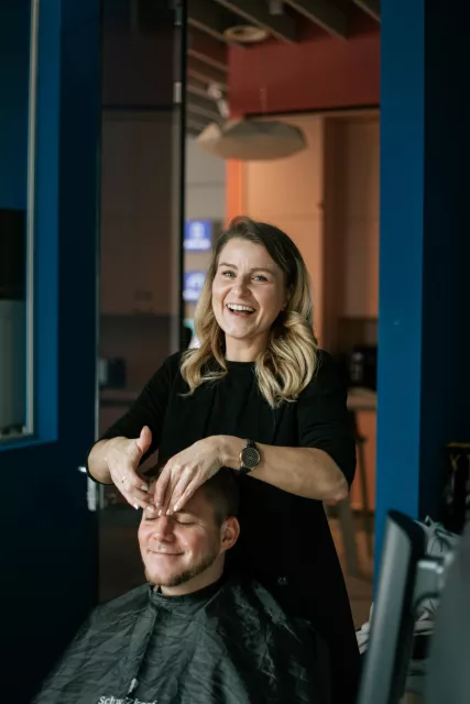 blonde frau lacht in die kamera, während sie einen mann vor sich im gesicht schminkt.