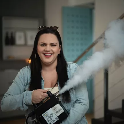 Kabarettistin Ina Jovanovic lacht in die Kamera und hält eine Nebelmaschine in der Hand, die Nebel versprüht.